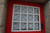 A window set in a red wooden frame, featuring a grid pattern with white lace curtains in each section. The window is bordered by textured stone walls, and an ornate black metal handle is visible on the right side.