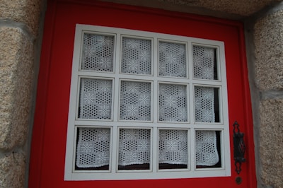 A window set in a red wooden frame, featuring a grid pattern with white lace curtains in each section. The window is bordered by textured stone walls, and an ornate black metal handle is visible on the right side.