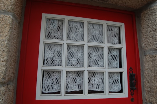 A window set in a red wooden frame, featuring a grid pattern with white lace curtains in each section. The window is bordered by textured stone walls, and an ornate black metal handle is visible on the right side.