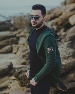 man in green jacket and black sunglasses standing on rocky shore during daytime