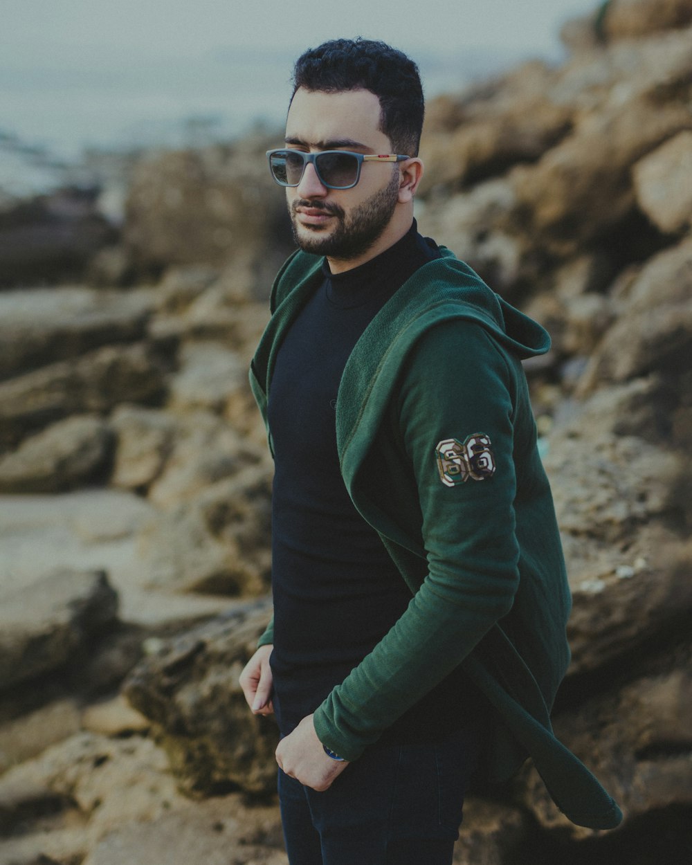 man in green jacket and black sunglasses standing on rocky shore during daytime