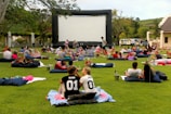 A large outdoor event with people gathered on a green lawn, sitting or lying on blankets and cushions, facing a big inflatable movie screen. The setting appears relaxed and social, with groups of people casually interacting. Trees and a building are seen in the background, and there are various items like baskets and coolers scattered around.