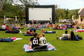 A large outdoor event with people gathered on a green lawn, sitting or lying on blankets and cushions, facing a big inflatable movie screen. The setting appears relaxed and social, with groups of people casually interacting. Trees and a building are seen in the background, and there are various items like baskets and coolers scattered around.