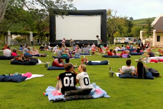 A large outdoor event with people gathered on a green lawn, sitting or lying on blankets and cushions, facing a big inflatable movie screen. The setting appears relaxed and social, with groups of people casually interacting. Trees and a building are seen in the background, and there are various items like baskets and coolers scattered around.