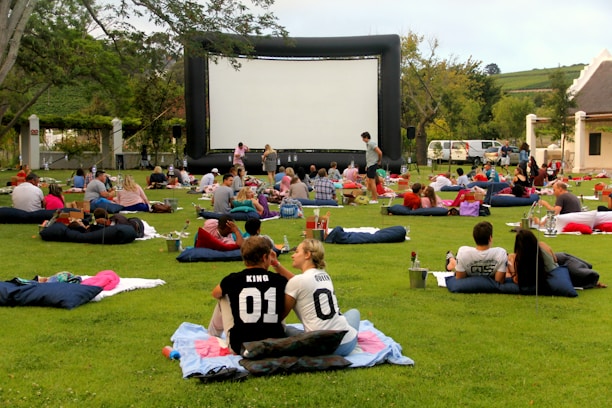 A large outdoor event with people gathered on a green lawn, sitting or lying on blankets and cushions, facing a big inflatable movie screen. The setting appears relaxed and social, with groups of people casually interacting. Trees and a building are seen in the background, and there are various items like baskets and coolers scattered around.