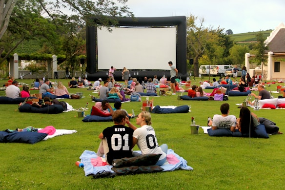 A large outdoor event with people gathered on a green lawn, sitting or lying on blankets and cushions, facing a big inflatable movie screen. The setting appears relaxed and social, with groups of people casually interacting. Trees and a building are seen in the background, and there are various items like baskets and coolers scattered around.