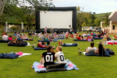 A large outdoor event with people gathered on a green lawn, sitting or lying on blankets and cushions, facing a big inflatable movie screen. The setting appears relaxed and social, with groups of people casually interacting. Trees and a building are seen in the background, and there are various items like baskets and coolers scattered around.