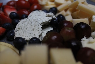Various types of cheese, grapes, blueberries, and sliced fruits are arranged together, creating a rich and textured assortment. The cheese includes a herbed variety, adding a speckled appearance. The fruits provide a contrast of colors and shapes.