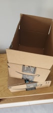 Three cardboard boxes with Amazon Prime branding are stacked on top of a wooden surface. The top box is open, revealing its interior. A light gray wall serves as the background.