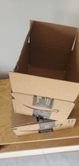 Three cardboard boxes with Amazon Prime branding are stacked on top of a wooden surface. The top box is open, revealing its interior. A light gray wall serves as the background.