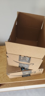 Three cardboard boxes with Amazon Prime branding are stacked on top of a wooden surface. The top box is open, revealing its interior. A light gray wall serves as the background.
