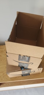 Three cardboard boxes with Amazon Prime branding are stacked on top of a wooden surface. The top box is open, revealing its interior. A light gray wall serves as the background.