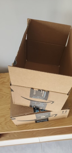 Three cardboard boxes with Amazon Prime branding are stacked on top of a wooden surface. The top box is open, revealing its interior. A light gray wall serves as the background.