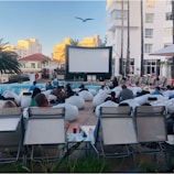 A group of people is gathered outdoors next to a pool, sitting on bean bags and lounge chairs, facing a large projection screen. The setting suggests an outdoor movie screening event. There are tall palm trees, buildings in the background, and a seagull flying above. The atmosphere appears casual and relaxed.