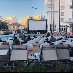 A group of people is gathered outdoors next to a pool, sitting on bean bags and lounge chairs, facing a large projection screen. The setting suggests an outdoor movie screening event. There are tall palm trees, buildings in the background, and a seagull flying above. The atmosphere appears casual and relaxed.