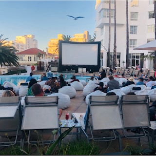 A group of people is gathered outdoors next to a pool, sitting on bean bags and lounge chairs, facing a large projection screen. The setting suggests an outdoor movie screening event. There are tall palm trees, buildings in the background, and a seagull flying above. The atmosphere appears casual and relaxed.