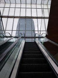 An escalator leads upwards towards large glass windows that reveal a modern skyscraper beyond. The architectural lines are sleek and the glass panels create a grid pattern.