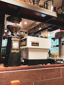 A coffee shop counter featuring a variety of coffee-making equipment, including a grinder and an espresso machine labeled 'Passmar'. The background includes shelves with jars and coffee accessories, and there's a warm ambient lighting.