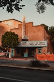 A vintage art deco theater with a marquee sign displaying the words 'Movies are forever, we'll see you soon'. The building is made of brick with teal blue accents on the door and windows. There are trees on either side of the building and an orange barrier on the sidewalk. The sky appears clear, suggesting it is either early morning or late afternoon.
