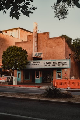 A vintage art deco theater with a marquee sign displaying the words 'Movies are forever, we'll see you soon'. The building is made of brick with teal blue accents on the door and windows. There are trees on either side of the building and an orange barrier on the sidewalk. The sky appears clear, suggesting it is either early morning or late afternoon.