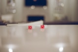 Two red dice with white dots are resting on a smooth, reflective surface. The background is blurred, giving focus to the dice in the foreground. The lighting is soft, which creates a serene and calm atmosphere around the dice.