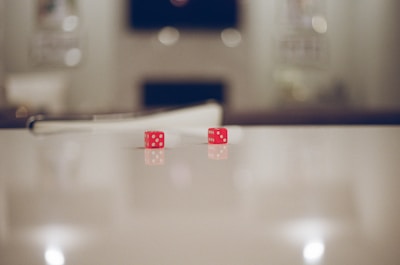 Two red dice with white dots are resting on a smooth, reflective surface. The background is blurred, giving focus to the dice in the foreground. The lighting is soft, which creates a serene and calm atmosphere around the dice.