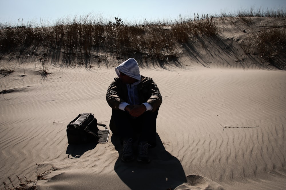 person in black hoodie and gray knit cap sitting on brown sand during daytime