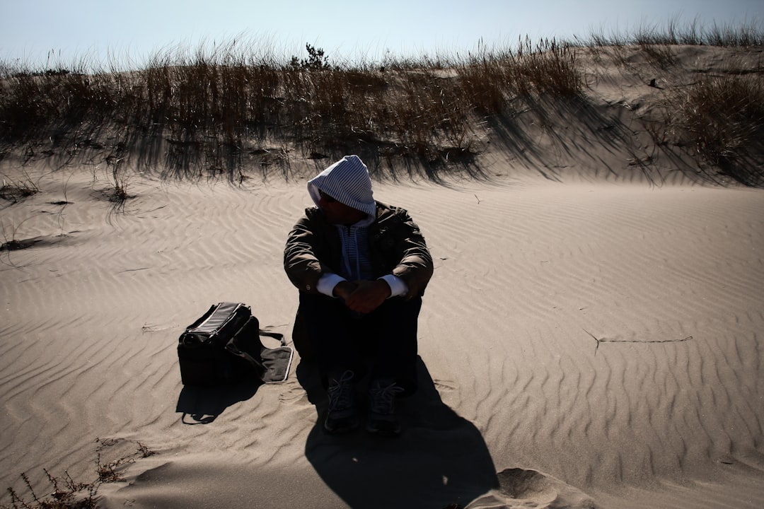 person in black hoodie and gray knit cap sitting on brown sand during daytime