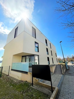 A modern, angular building with clean lines and a flat roof. The facade is cream-colored with large, dark-framed windows. Sunlight is illuminating the building from above, casting a bright glow. A dark sign stands in front of the building, and a paved sidewalk runs alongside.