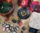 A variety of handmade ceramic items are displayed on a light brown surface. These include colorful plates with floral designs, a large green bowl, several decorative pieces shaped like flowers, leaves, and other nature motifs. Prices are marked with numbers written on small pieces of paper placed next to some items.