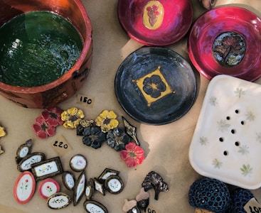 A variety of handmade ceramic items are displayed on a light brown surface. These include colorful plates with floral designs, a large green bowl, several decorative pieces shaped like flowers, leaves, and other nature motifs. Prices are marked with numbers written on small pieces of paper placed next to some items.