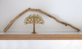 A minimalist wooden shelf displays a decorative wooden tree positioned beneath an arched branch. The background is a plain white wall, emphasizing the natural elements and simple design.