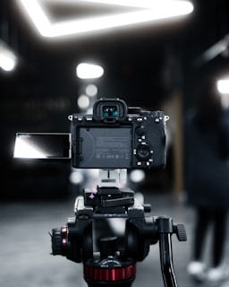 A professional camera mounted on a tripod, focused on the back view of the camera with an extended flip-out screen. The setting appears to be indoors with a blurred background and bright, geometric ceiling lights.