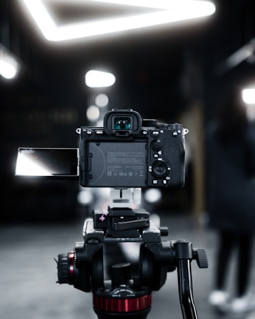 A professional camera mounted on a tripod, focused on the back view of the camera with an extended flip-out screen. The setting appears to be indoors with a blurred background and bright, geometric ceiling lights.