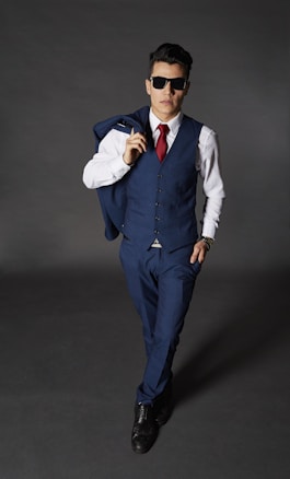 A person is dressed in a tailored blue suit with a white shirt and a red tie, accessorized with sunglasses and a watch, posing confidently against a dark background. They hold a matching blue jacket over the shoulder and wear black shoes.