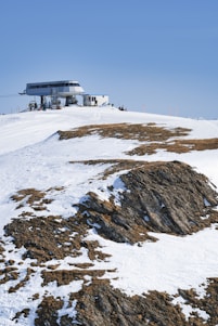 A snow-covered mountain landscape with patches of exposed rock, featuring a modern cable car station situated at the crest of the hill. The clear blue sky provides a backdrop and a few people are visible near the station, likely resting or preparing for skiing or snowboarding.