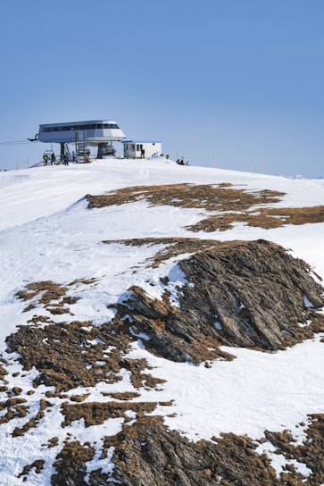 A snow-covered mountain landscape with patches of exposed rock, featuring a modern cable car station situated at the crest of the hill. The clear blue sky provides a backdrop and a few people are visible near the station, likely resting or preparing for skiing or snowboarding.