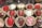 An assortment of decorated strawberries placed in brown paper cupcake holders. Some strawberries are coated with chocolate and decorated with red and white icing, while others are covered with sugar crystals. The strawberries are arranged in neat rows on a dark marble surface.