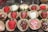 An assortment of decorated strawberries placed in brown paper cupcake holders. Some strawberries are coated with chocolate and decorated with red and white icing, while others are covered with sugar crystals. The strawberries are arranged in neat rows on a dark marble surface.
