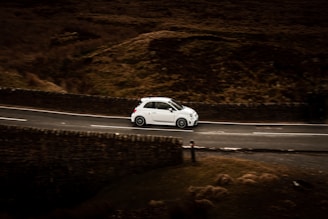 A small white car is driving along a winding road, which is bordered by stone walls. The surrounding landscape is rugged and appears to be a rural or countryside area with earthy, brown tones.