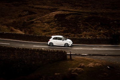 A small white car is driving along a winding road, which is bordered by stone walls. The surrounding landscape is rugged and appears to be a rural or countryside area with earthy, brown tones.
