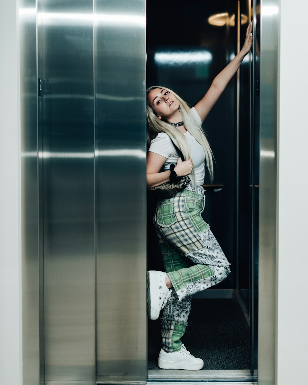 woman in green and white plaid dress shirt and black pants leaning on gray wall