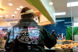 A person wearing a dark hoodie with the Krispy Kreme logo stands inside a bakery or store, overseeing a machine with freshly prepared donuts. The setting has a warm, indoor lighting with several hanging lamps visible. In the background, there are staff members working and various store items.