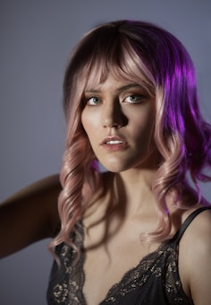 A close-up portrait of a woman with pink wavy hair and bangs, illuminated by soft lighting. She wears a dark, lace-trimmed camisole and gazes directly at the camera. The background is a muted gradient, enhancing the focus on her facial features and hair.