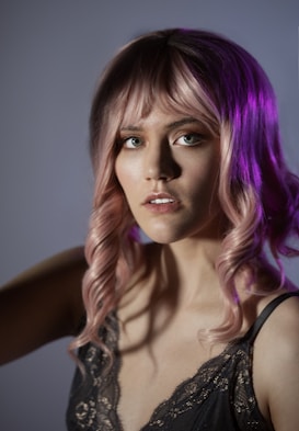 A close-up portrait of a woman with pink wavy hair and bangs, illuminated by soft lighting. She wears a dark, lace-trimmed camisole and gazes directly at the camera. The background is a muted gradient, enhancing the focus on her facial features and hair.