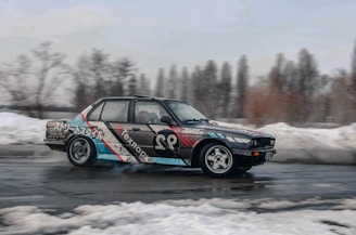 A modified car is drifting on a wet asphalt surface. The vehicle has a colorful design featuring various graphics and text on its body. In the background, snow-covered ground and leafless trees suggest a cold and wintry setting.
