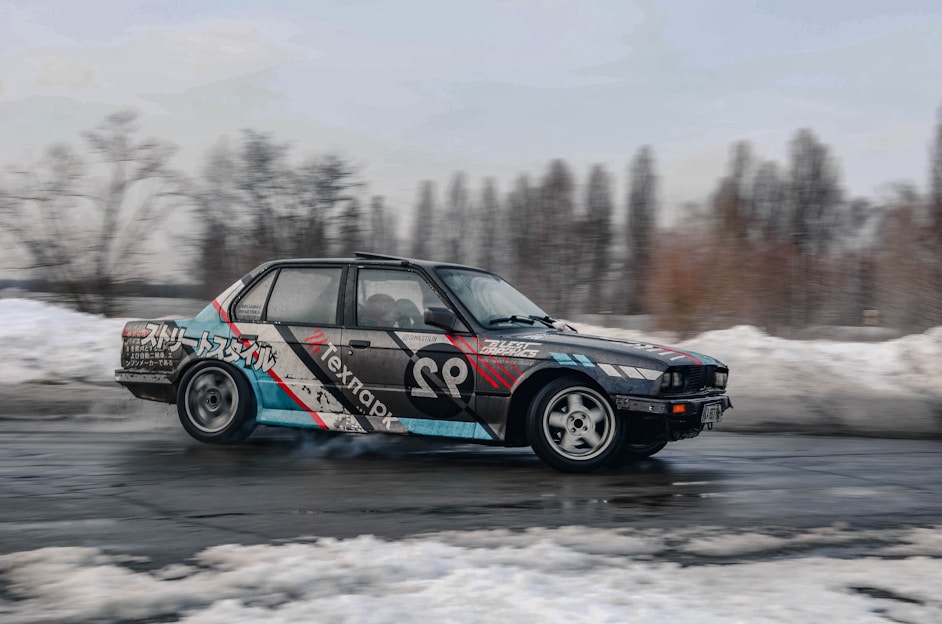 A modified car is drifting on a wet asphalt surface. The vehicle has a colorful design featuring various graphics and text on its body. In the background, snow-covered ground and leafless trees suggest a cold and wintry setting.
