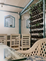 The image depicts a cozy indoor setting with a mix of natural and modern elements. There are white wooden chairs arranged around a table, and a glass-topped table in the foreground. A wall display features decorative blue striped patterns. A vertical garden with multiple small planters showcasing green plants is mounted on a metal frame. The atmosphere is calm and inviting.