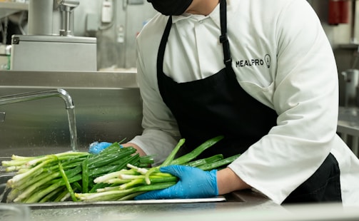 A person wearing a white chef's coat and black apron, with gloves on, is washing green onions under a running tap in a commercial kitchen setting. The setting includes a stainless steel sink and kitchen equipment in the background.