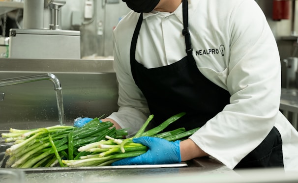 A person wearing a white chef's coat and black apron, with gloves on, is washing green onions under a running tap in a commercial kitchen setting. The setting includes a stainless steel sink and kitchen equipment in the background.